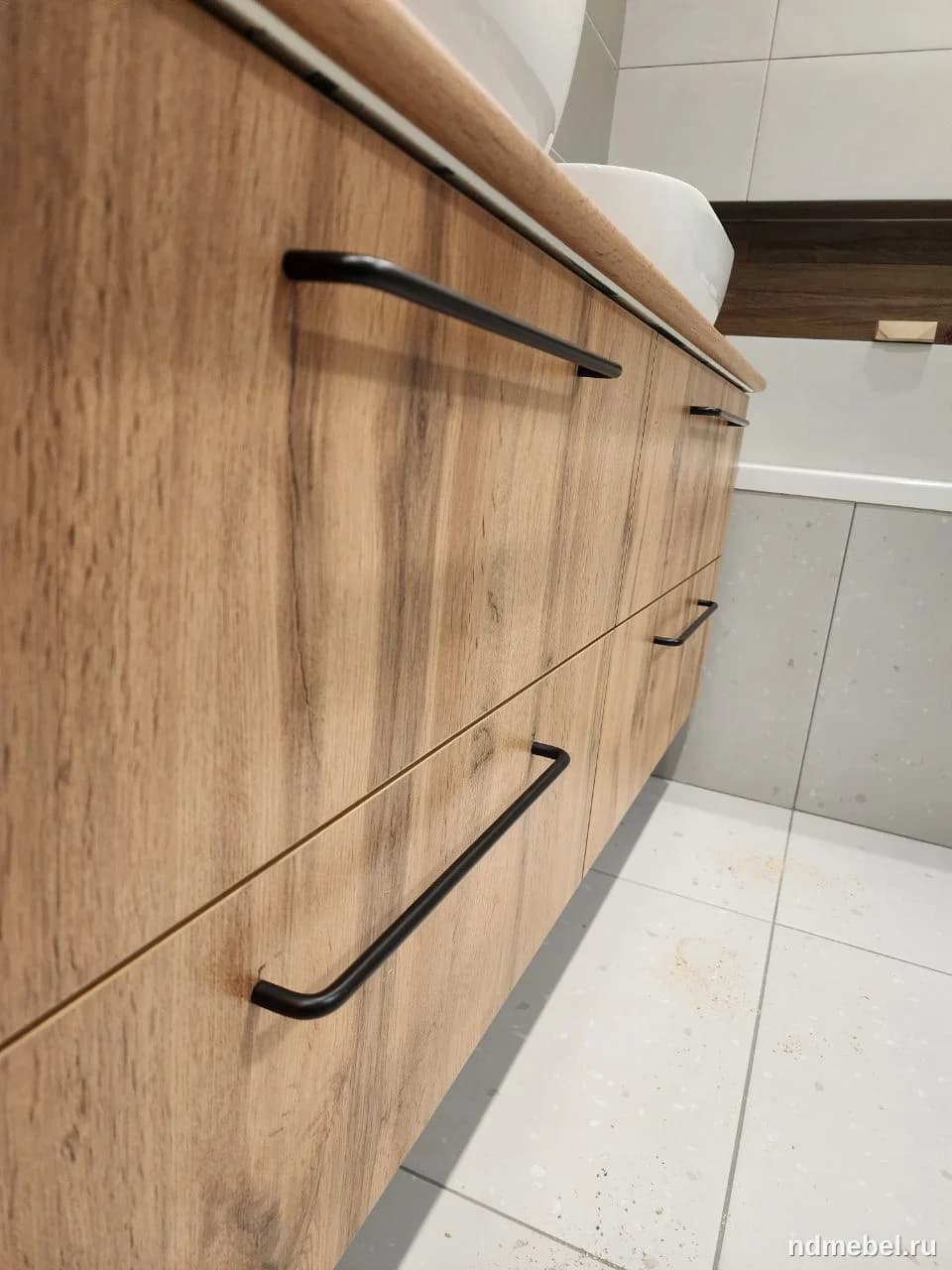 Тумба с двумя выдвижными ящиками, выполненная под натуральное дерево светлого оттенка с выраженной текстурой. Мебель дополнена современными длинными черными матовыми ручками, что придает ей минималистичный и актуальный вид.