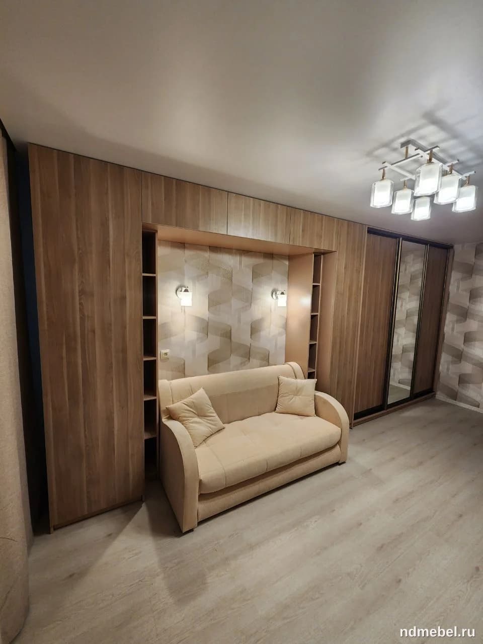Встроенный мебельный гарнитур из светлого дерева с нишей для дивана и встроенным шкафом-купе, создающий функциональное и теплое пространство в современном стиле. Центральная зона ниши отделана геометрическими панелями и оборудована подсветкой, дополняя скромный бежевый диван.