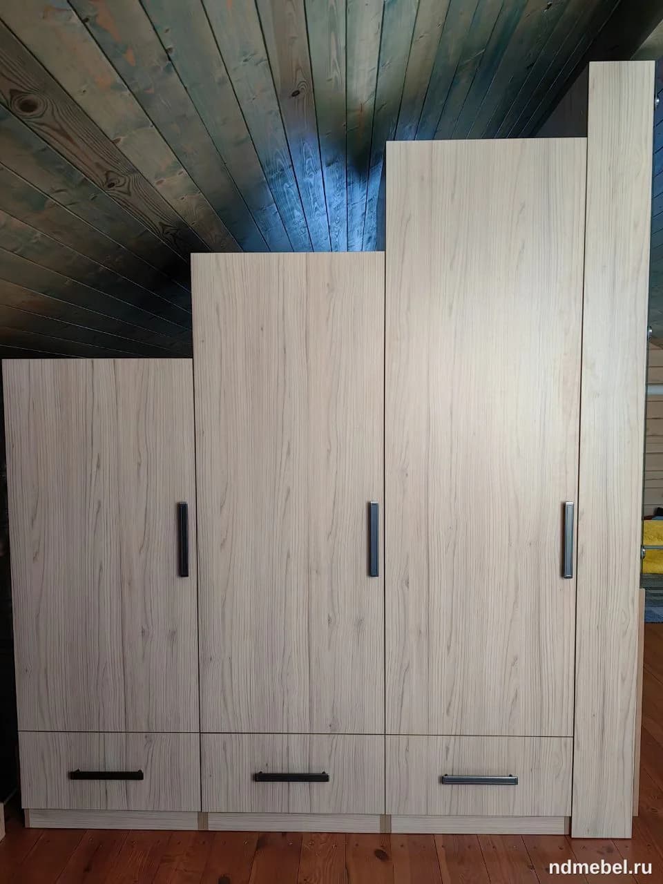 Это модульный или секционный шкаф светлого древесного (возможно, дубового) цвета с матовыми черными вертикальными ручками на дверцах и горизонтальными на нижних ящиках. Мебель выполнена в современном минималистичном стиле и имеет асимметричную высоту секций, что может быть обусловлено особенностями помещения (например, скошенным потолком, как видно на фоне).