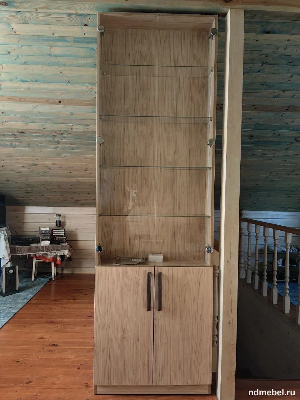 Высокий витринный шкаф с комбинированными секциями: верхняя часть со стеклянными полками и прозрачной дверцей, нижняя — с глухими деревянными дверцами. Мебель выполнена из светлого дерева с выраженной текстурой, что придает ей современный, но теплый вид.