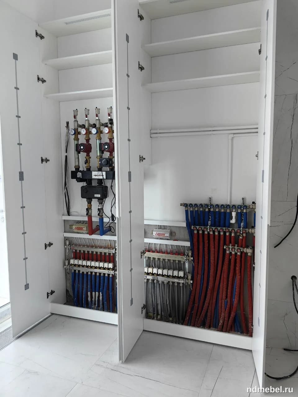 Встроенный белый шкаф-колонна, выполненный в современном минималистичном стиле, предназначен для маскировки инженерных коммуникаций, включая коллекторы теплого пола и запорную арматуру. Мебель имеет глухие фасады и внутренние полки, что обеспечивает аккуратный и чистый вид помещения.