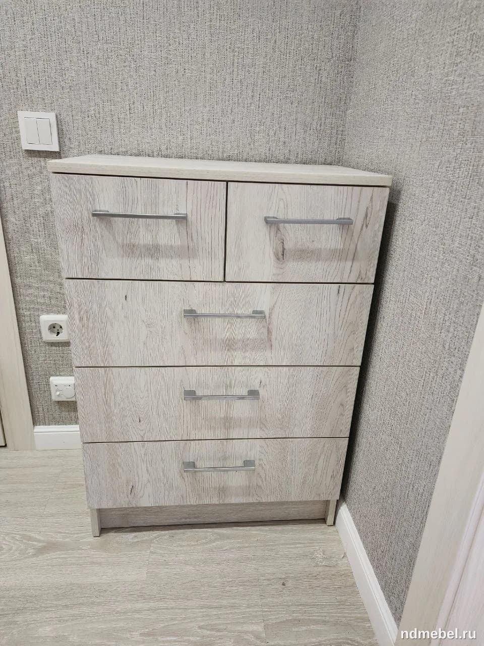 Тумба (комод) с пятью ящиками (два маленьких сверху и три больших снизу) выполнена из материала, имитирующего светлое, выбеленное дерево. Мебель имеет современный простой стиль с минималистичными металлическими ручками.