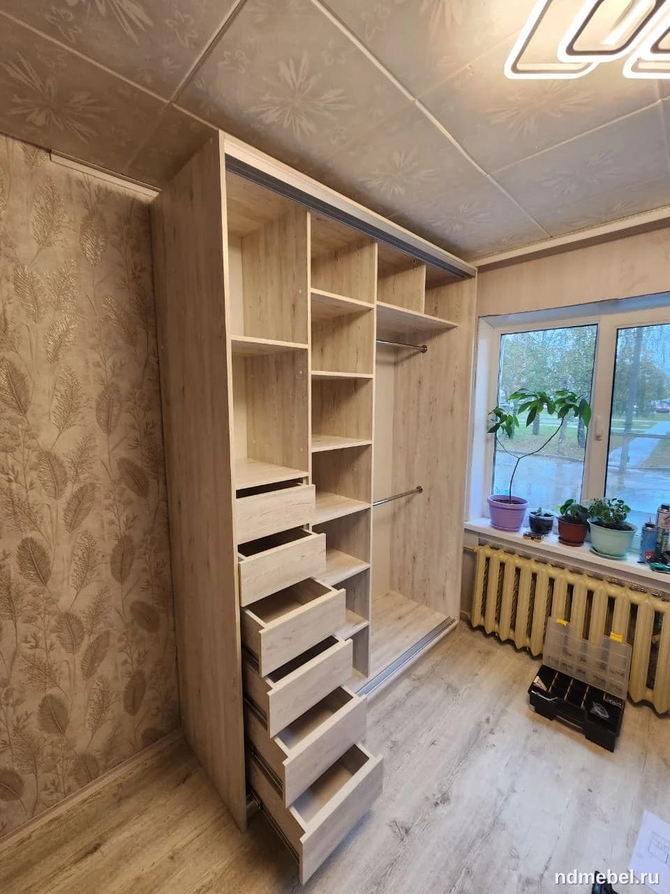 Встроенный шкаф-купе с раздвижными дверцами и открытой секцией, демонстрирующий внутреннее наполнение из светлого ламинированного ДСП с текстурой под дерево. Мебель выполнена в современном минималистичном стиле и имеет множество полок и выдвижных ящиков для хранения.