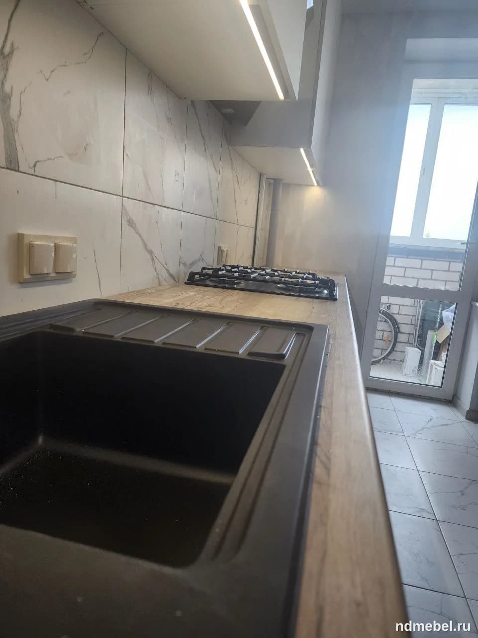 На изображении показана часть кухонной столешницы из светлого дерева с интегрированной черной мойкой и варочной панелью. Фартук выполнен из глянцевой белой плитки, имитирующей мрамор, что создает современный и светлый интерьер.