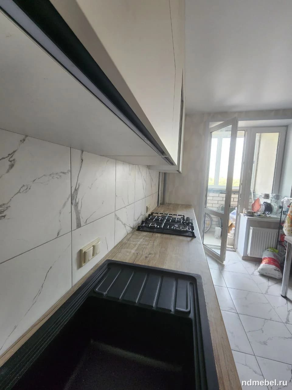 Современная кухонная столешница с текстурой светлого дерева, дополненная черной мойкой и двумя светлыми верхними шкафами с интегрированной подсветкой. Фартук выполнен из глянцевой плитки, имитирующей белый мрамор с серыми прожилками.