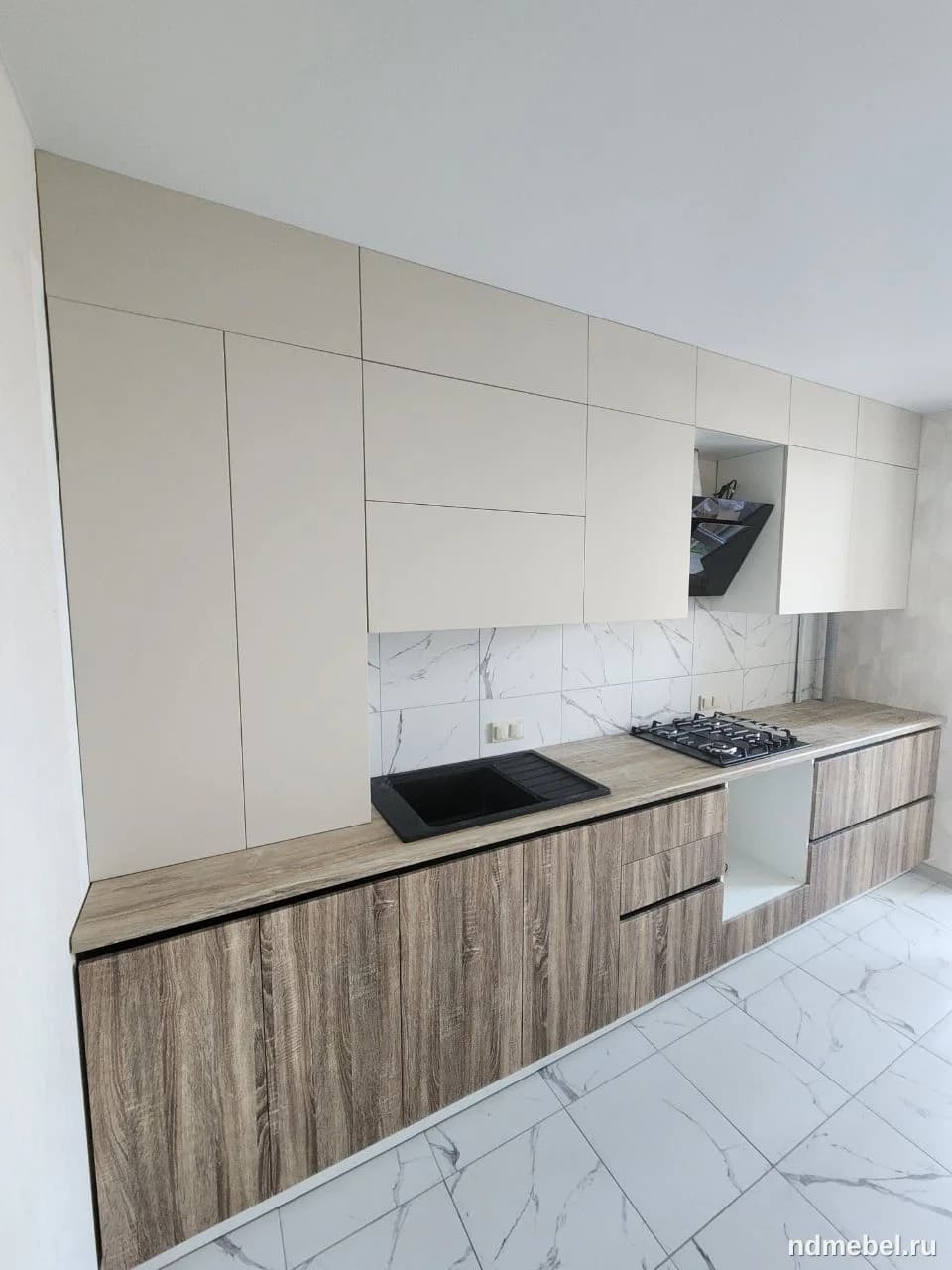 Это современная прямая кухонная гарнитура с нижними фасадами под светлое дерево и верхними гладкими, бежевыми или светло-серыми фасадами без ручек. Стиль минималистичный, с контрастом между текстурой дерева и гладкими матовыми поверхностями.