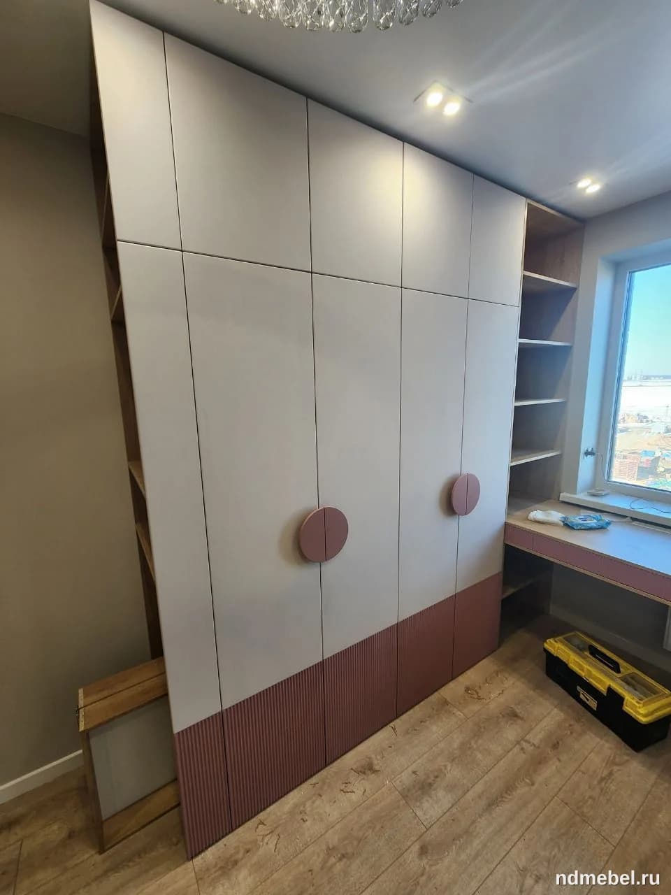 Это большой, встроенный шкаф-гардероб с современным дизайном, выполненный в двухцветной гамме: светло-серые/бежевые глянцевые фасады и нижняя часть с декоративными рифлеными панелями пыльно-розового цвета. Мебель дополнена круглыми ручками в тон нижней части и интегрирована с открытыми полками и рабочим местом у окна.