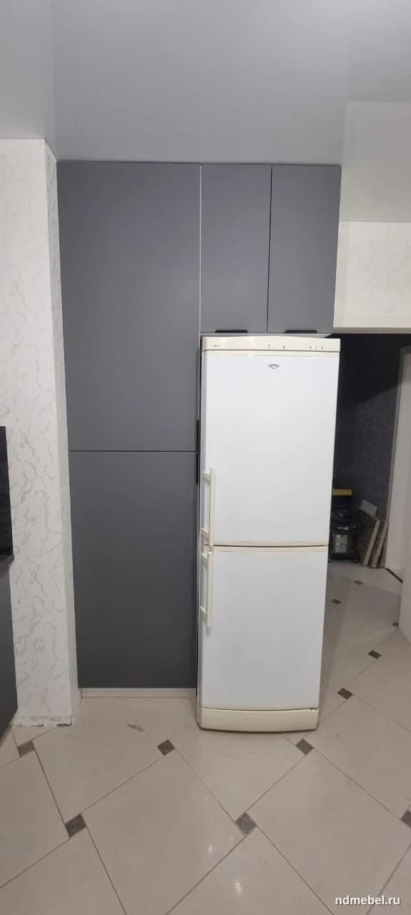 Встроенный кухонный шкаф или пенал, выполненный из матового материала темно-серого (графитового) цвета, имеет современный минималистичный стиль. Шкаф расположен рядом с отдельно стоящим белым двухкамерным холодильником.