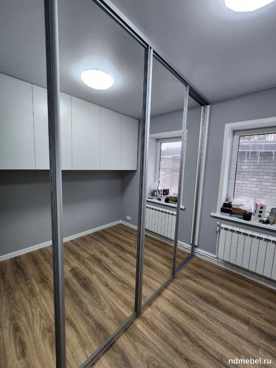 Встроенный шкаф-купе с зеркальными фасадами в алюминиевом профиле занимает целую стену, его отражающая поверхность визуально расширяет пространство небольшой комнаты с ламинатом под дерево.