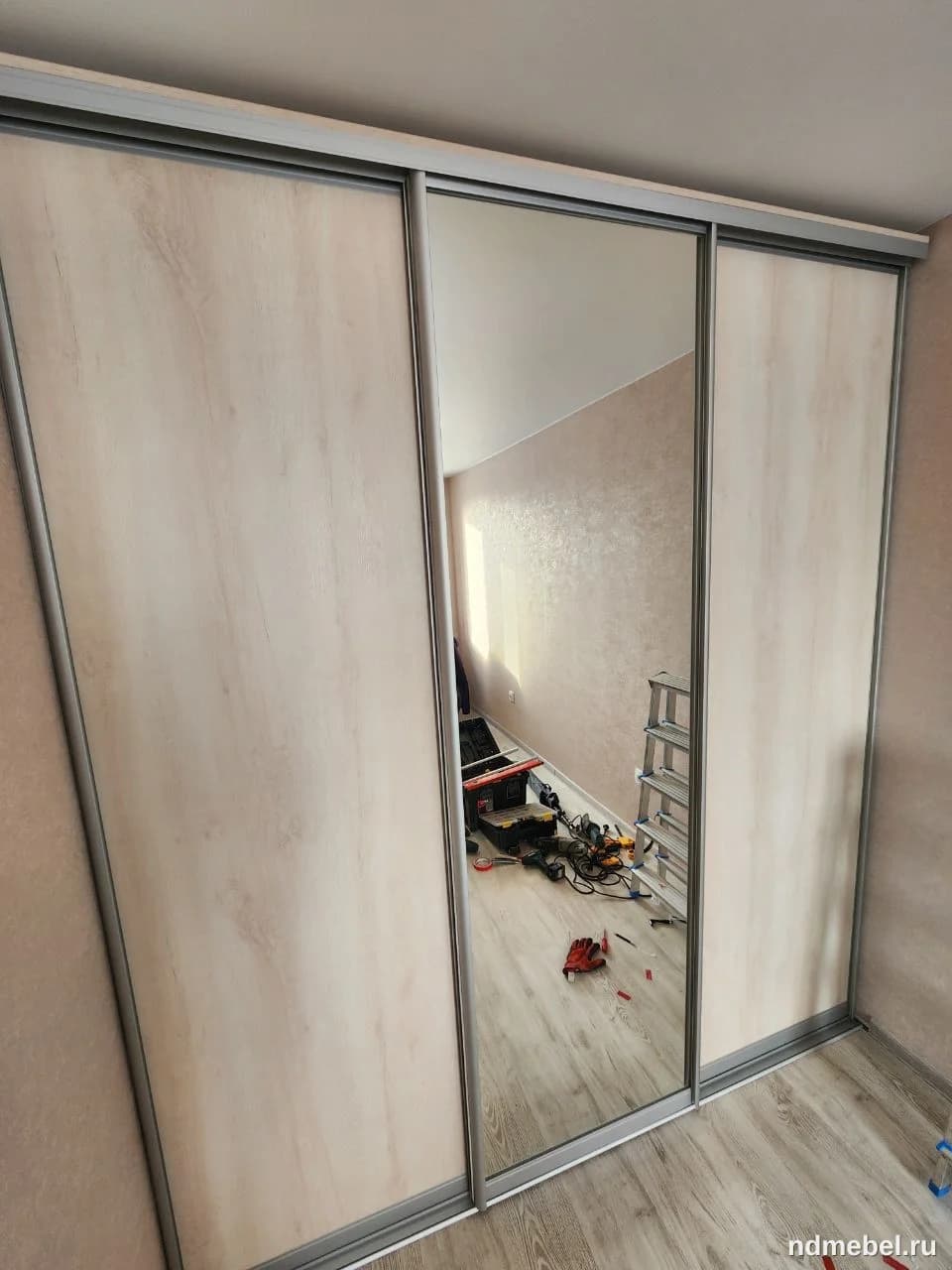 Встроенный или отдельно стоящий шкаф-купе с тремя раздвижными дверями, выполненный в современном стиле с фасадами из светлого ЛДСП под дерево и одной зеркальной вставкой по центру, обрамленными серебристым алюминиевым профилем. Мебель универсальна и подойдет для хранения вещей в спальне или прихожей.