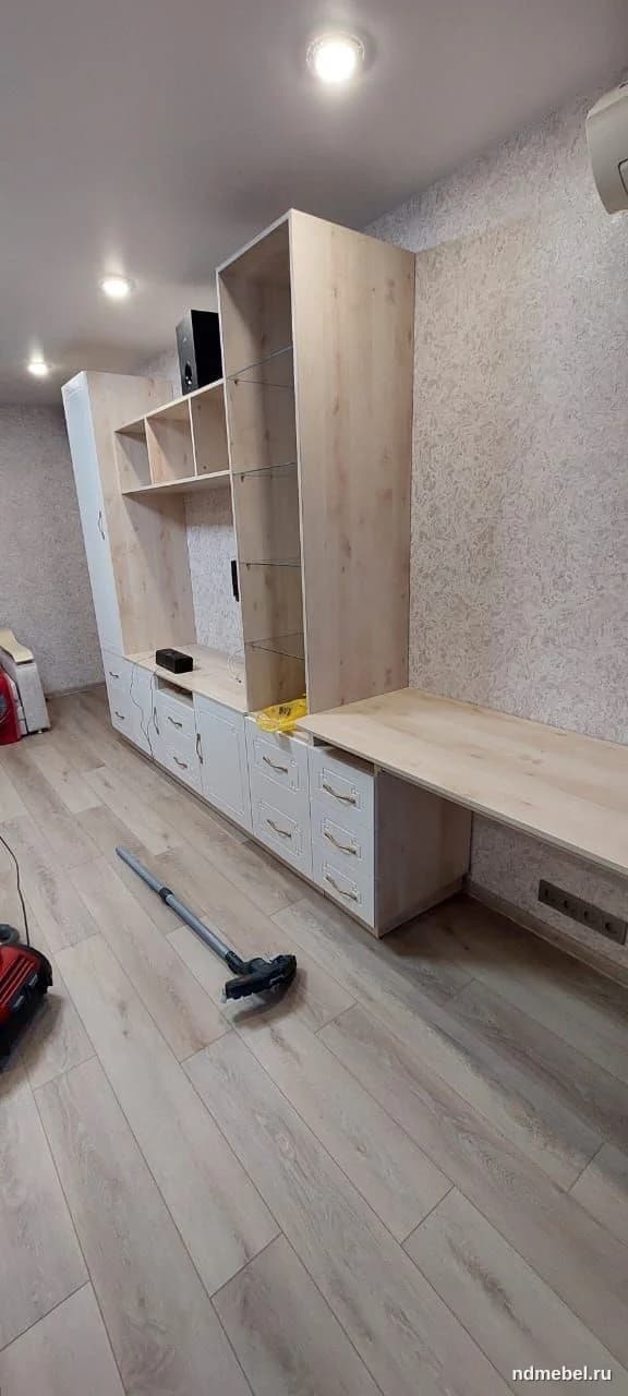 Светлая модульная стенка для гостиной, выполненная из ЛДСП с имитацией светлого дерева и белыми фасадами ящиков. Мебель выполнена в современном стиле с акцентом на функциональность и минималистичные светлые тона.