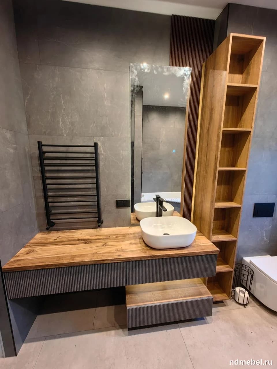 Современная подвесная тумба под раковину в стиле лофт/минимализм, выполненная из комбинации столешницы под натуральное дерево и темно-серого корпуса с вертикальными фрезерованными элементами. Комплект дополнен высоким открытым стеллажом из дерева и белой накладной раковиной.