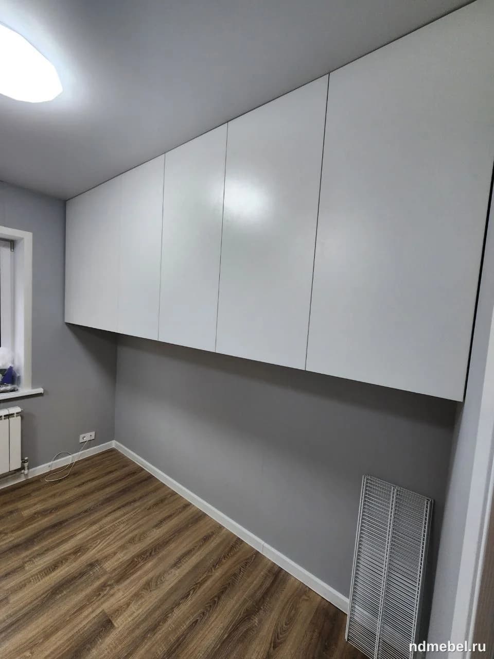 Встроенный навесной шкаф с глянцевыми белыми фасадами без ручек, выполненный в современном минималистичном стиле. Мебель занимает всю ширину стены над зоной, оставляя открытое пространство под собой.