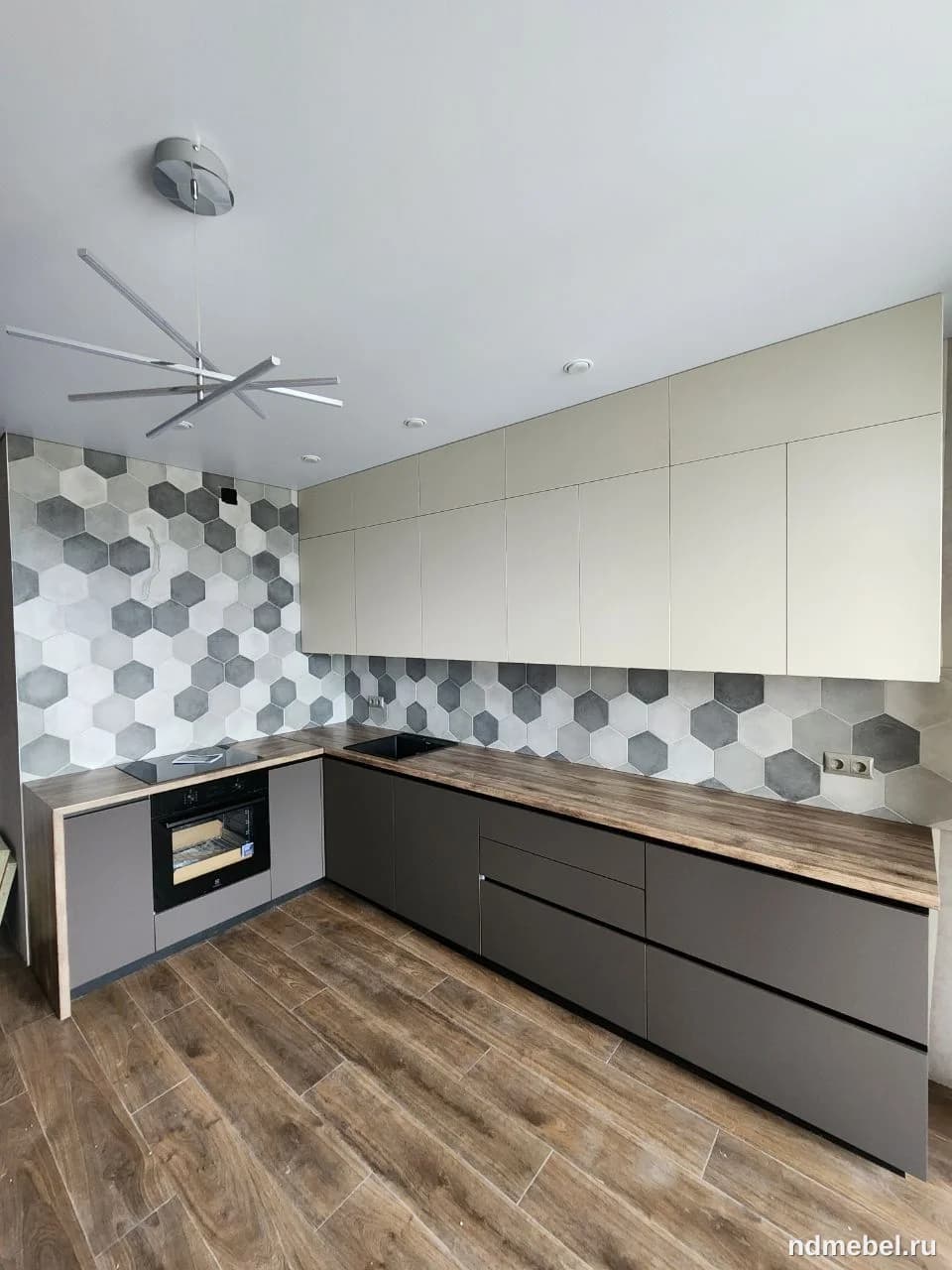 Современная угловая кухонная гарнитура с матовыми фасадами в темно-сером цвете и бежевыми верхними шкафами, дополненная столешницей под темное дерево и акцентным шестиугольным фартуком.