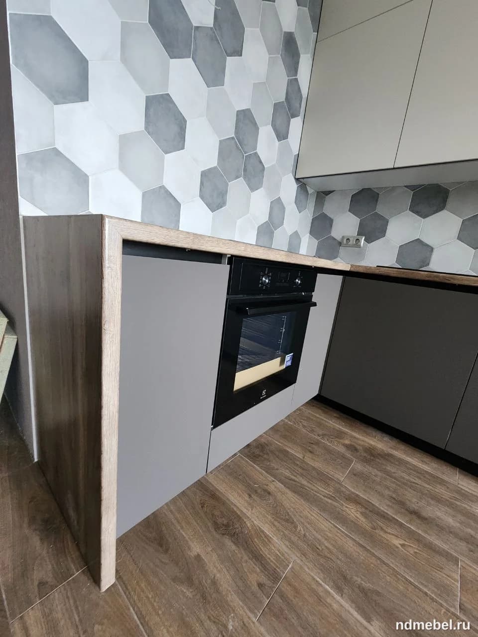 Современная кухонная мебель в минималистичном стиле, сочетающая фасады серого цвета с элементами под светлое дерево и встроенной черной техникой. Интерьер дополнен оригинальным шестиугольным фартуком в оттенках серого и белого.