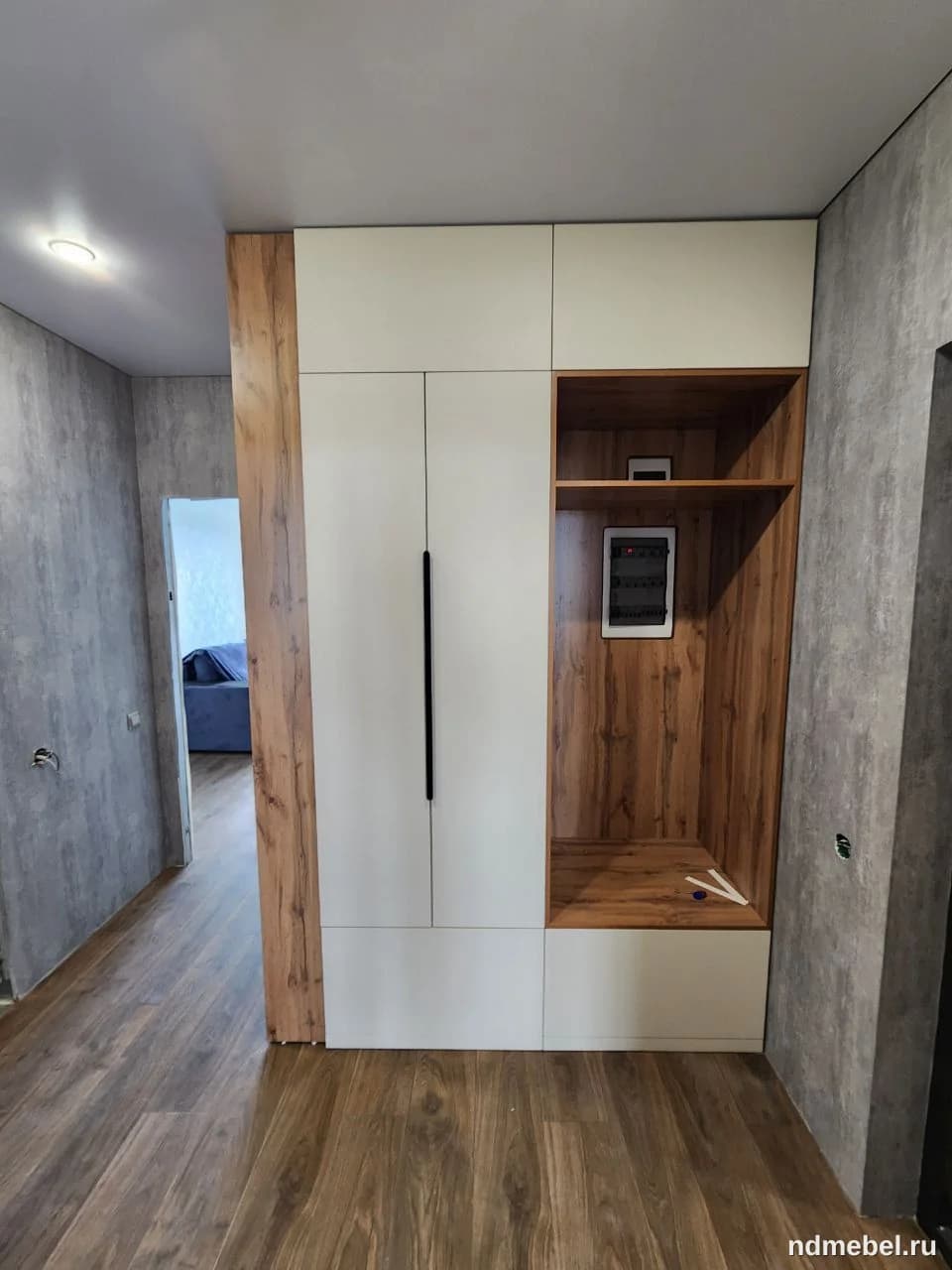 Современный высокий шкаф-пенал, сочетающий белые матовые фасады с текстурой темного дерева. Мебель имеет минималистичный дизайн с вертикальными черными ручками и открытыми нишами, что характерно для прихожей или малогабаритного жилого пространства.