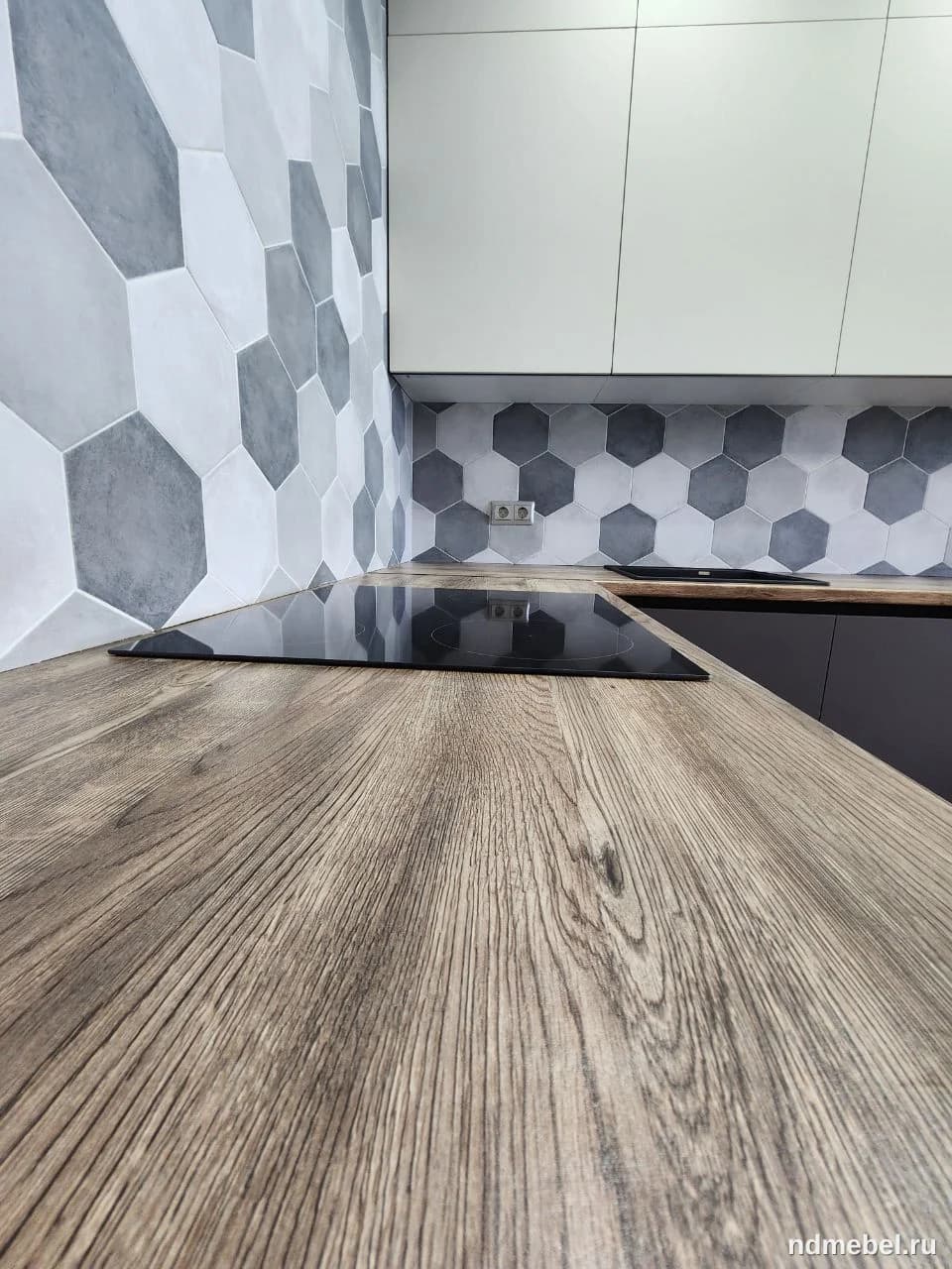 Современная кухонная столешница с текстурой под серо-коричневое дерево дополнена глянцевой черной варочной панелью и фартуком из шестиугольной плитки в оттенках серого и белого. Мебель выполнена в минималистичном современном стиле с акцентом на контраст текстур и цветов.