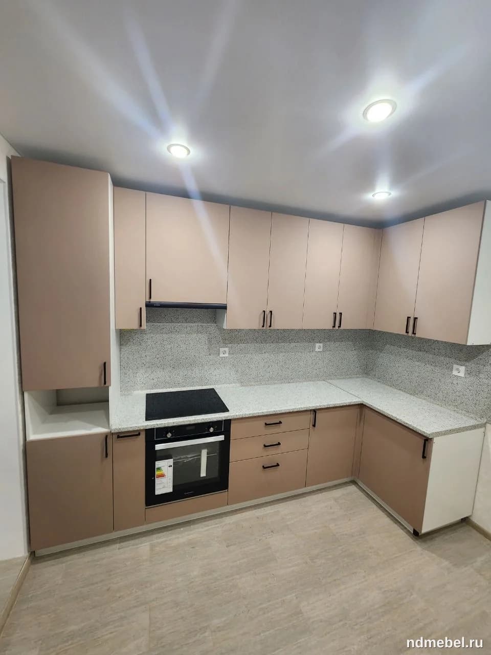 Это современный кухонный гарнитур, выполненный в минималистичном стиле с гладкими фасадами теплого пудрового или бежево-розового оттенка и светлой столешницей с мелким краплением. Мебель имеет лаконичные черные ручки и встраиваемую черную технику.