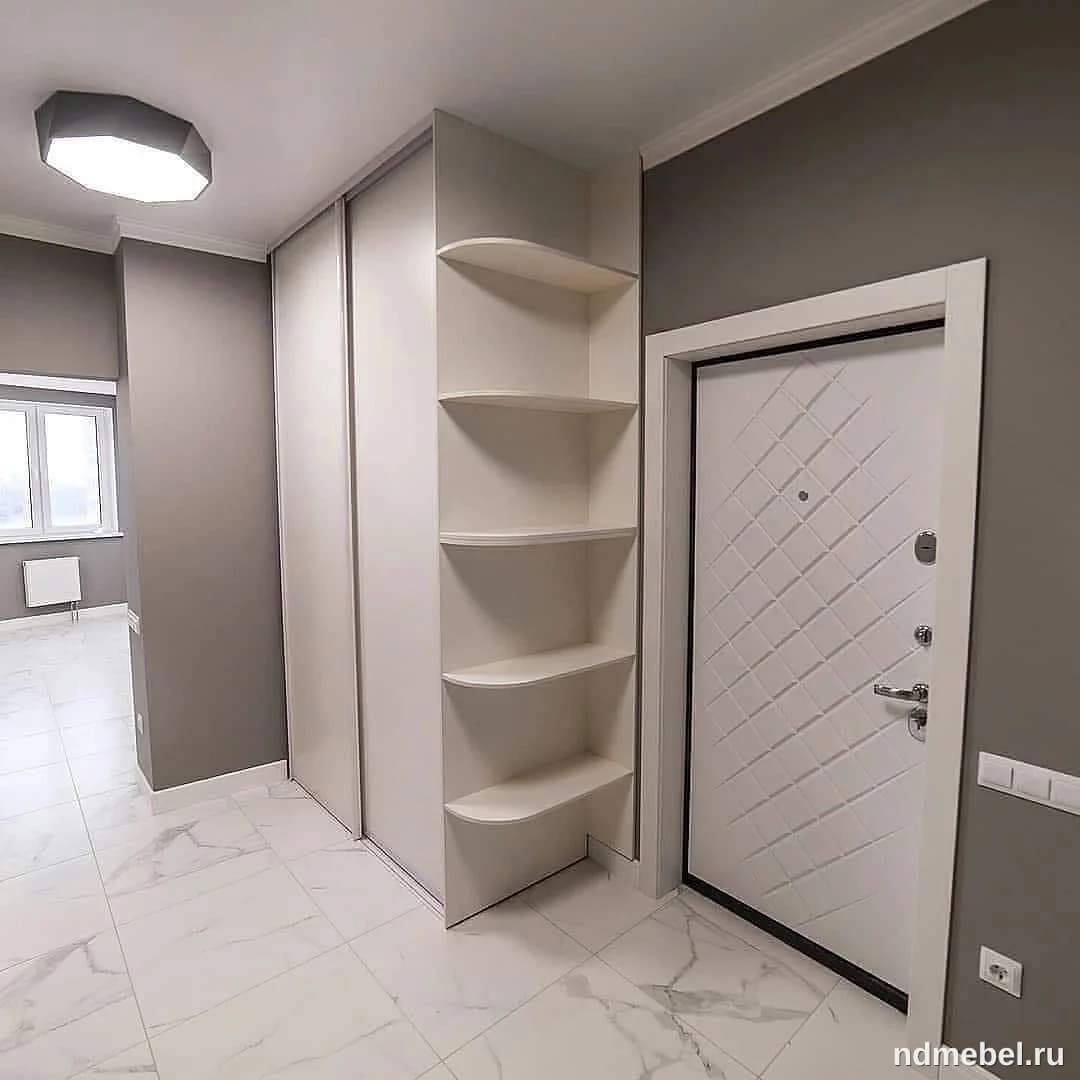 Встроенный шкаф-купе с белыми глянцевыми дверцами и открытыми угловыми полками, выполненный в современном минималистичном стиле. Мебель имеет светлый цвет и доминирует в сером интерьере прихожей с мраморным полом.