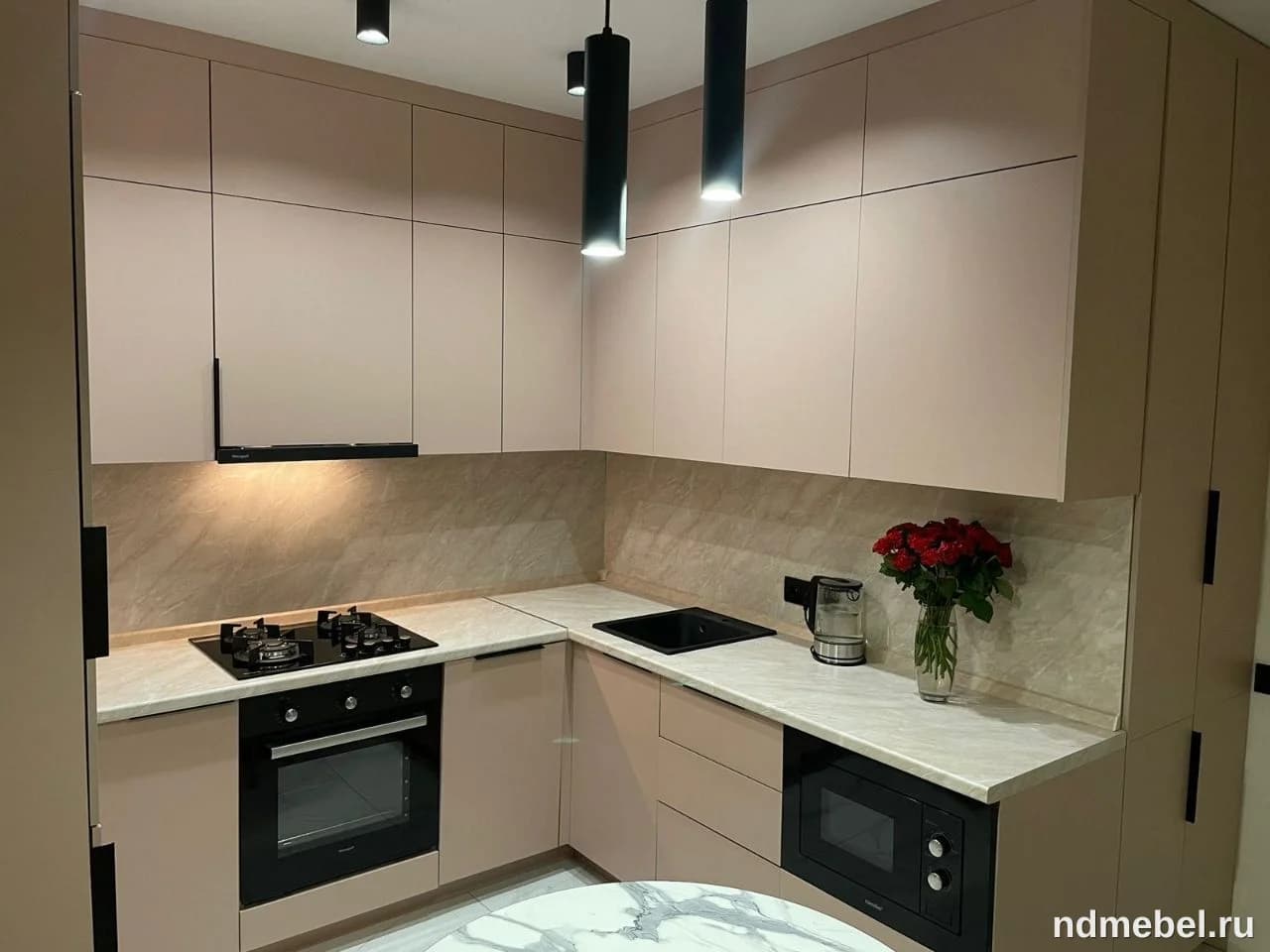 Современная угловая кухня с гладкими фасадами светло-розового (пудрового) цвета и светлой мраморной столешницей. Мебель выполнена в минималистичном стиле с черной встроенной техникой и вертикальными черными ручками.